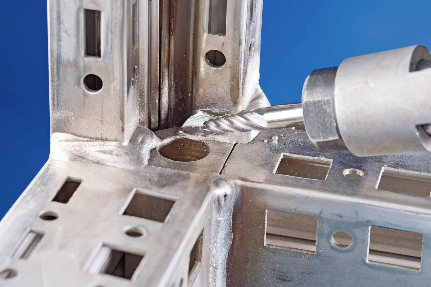 Wiertło metalowe obrabia precyzyjnie wykonany element metalowy z wieloma otworami i rowkami na niebieskim tle.