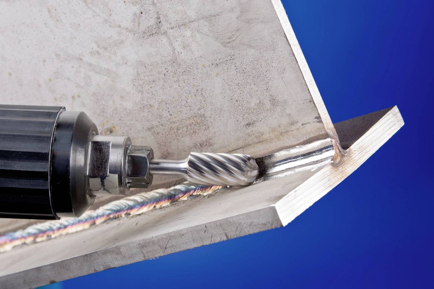 Zbliżenie spawu metalowego. Elektryczne narzędzie z obrotową tarczą obrabia spaw pod kątem. Tło w kolorze niebieskim.