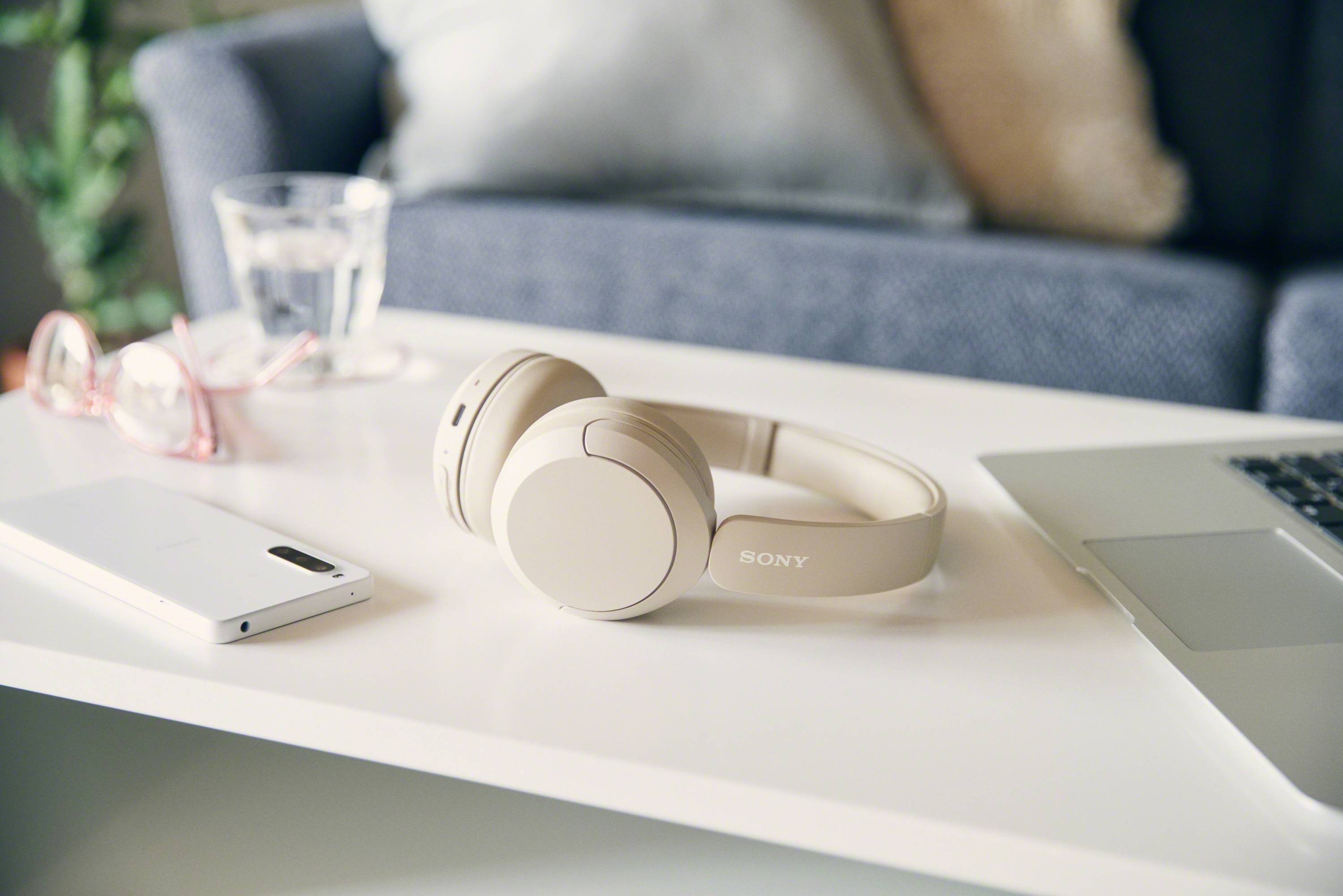 Bezprzewodowe słuchawki na białym stole obok laptopa i smartfona. W tle sofa i szklanka wody.