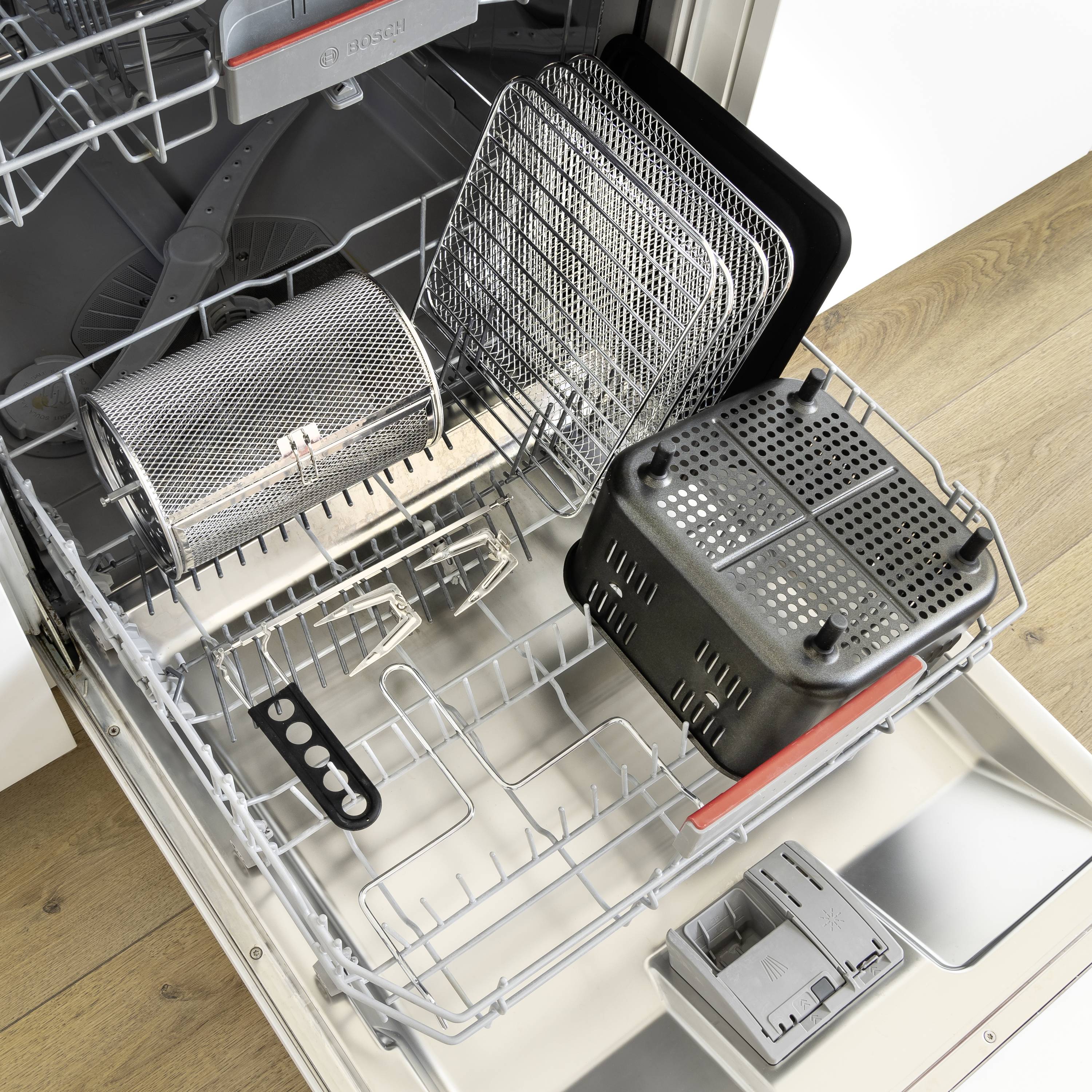 Zmywarka z otwartymi drzwiczkami, wyposażona w kilka metalowych koszy, tacę na sztućce oraz kosz filtracyjny w dolnej części.