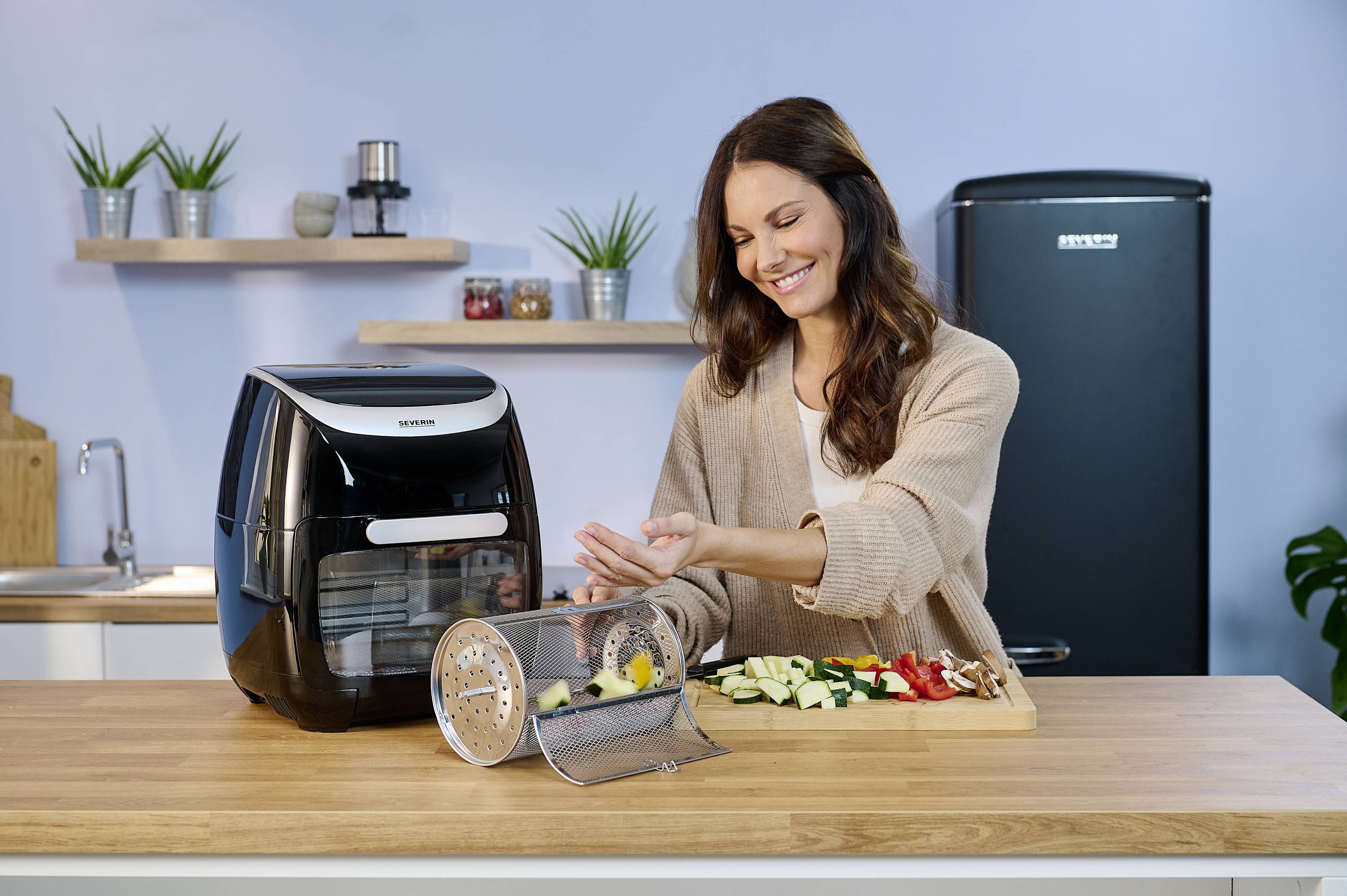 Kobieta przygotowuje warzywa w nowoczesnej kuchni. Obok niej na blacie stoi frytownica beztłuszczowa. W tle widać różne przybory kuchenne.