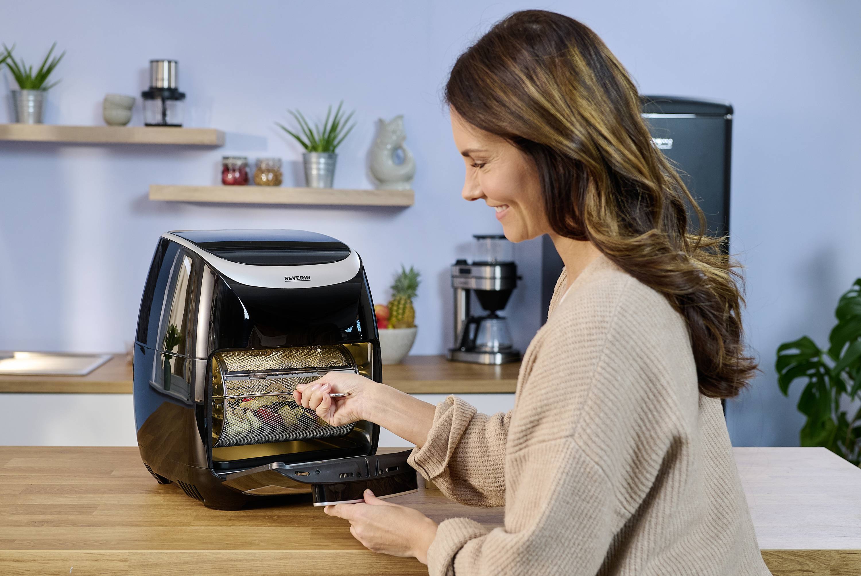 Kobieta używa air fryer w nowoczesnej kuchni. Wyjmuje jedzenie z urządzenia. W tle widać półki kuchenne i rośliny.