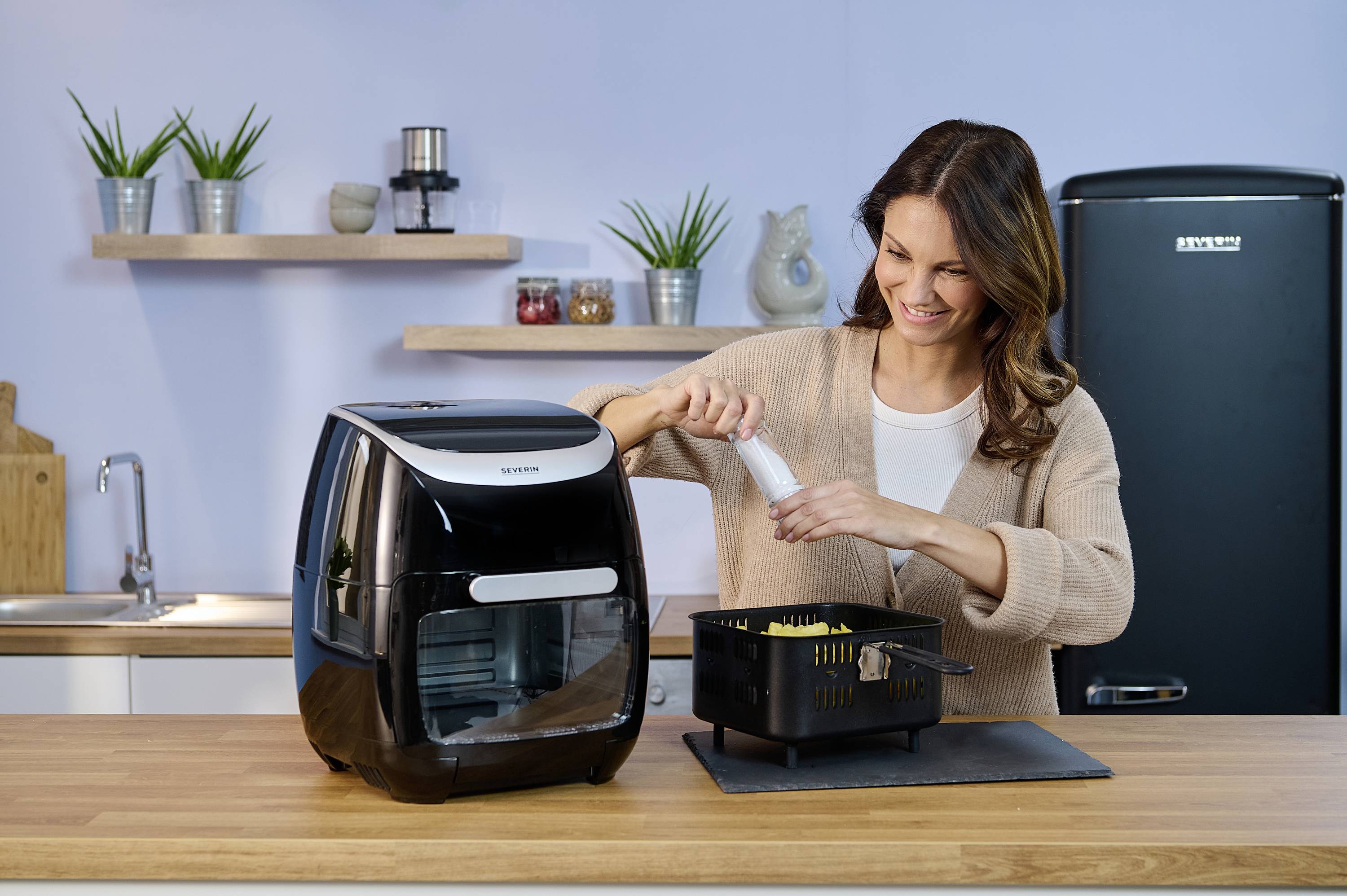 Kobieta w kuchni napełnia frytkownicę ziemniakami, uśmiechając się. W tle widać półki z roślinami i przyprawami. Scena przedstawia gotowanie w domu.