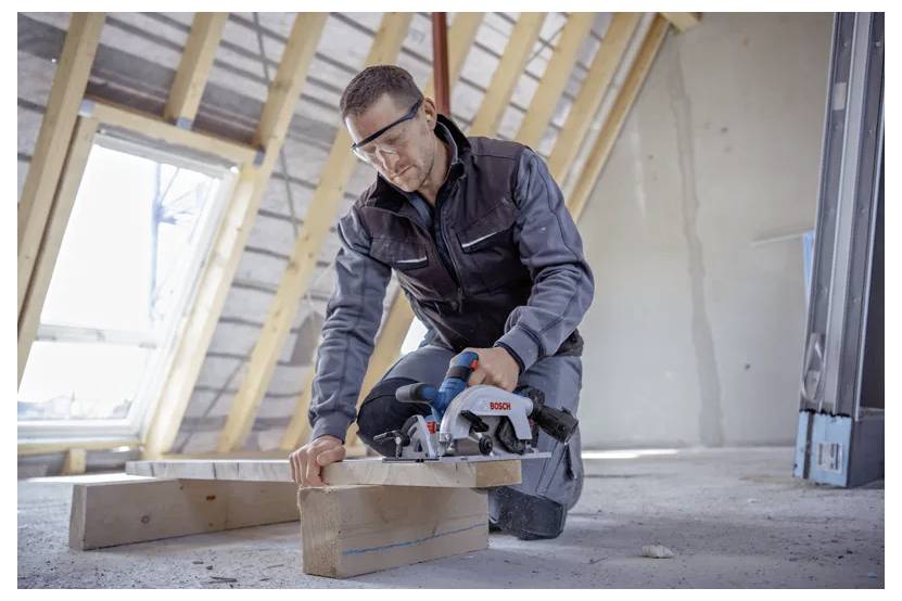 Osoba w okularach ochronnych przecina drewniane deski elektryczną piłą w nieukończonym poddaszu, otoczona nieosłoniętymi belkami.