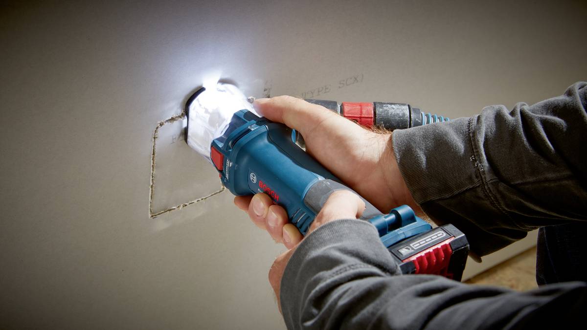 Osoba używa niebieskiego urządzenia elektrycznego, aby wyciąć prostokątny otwór w ścianie z płyty gipsowo-kartonowej.