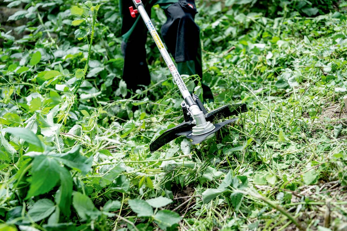 Osoba kosi gęste zarośla za pomocą podkaszarki w zielonym, porośniętym ogrodzie.