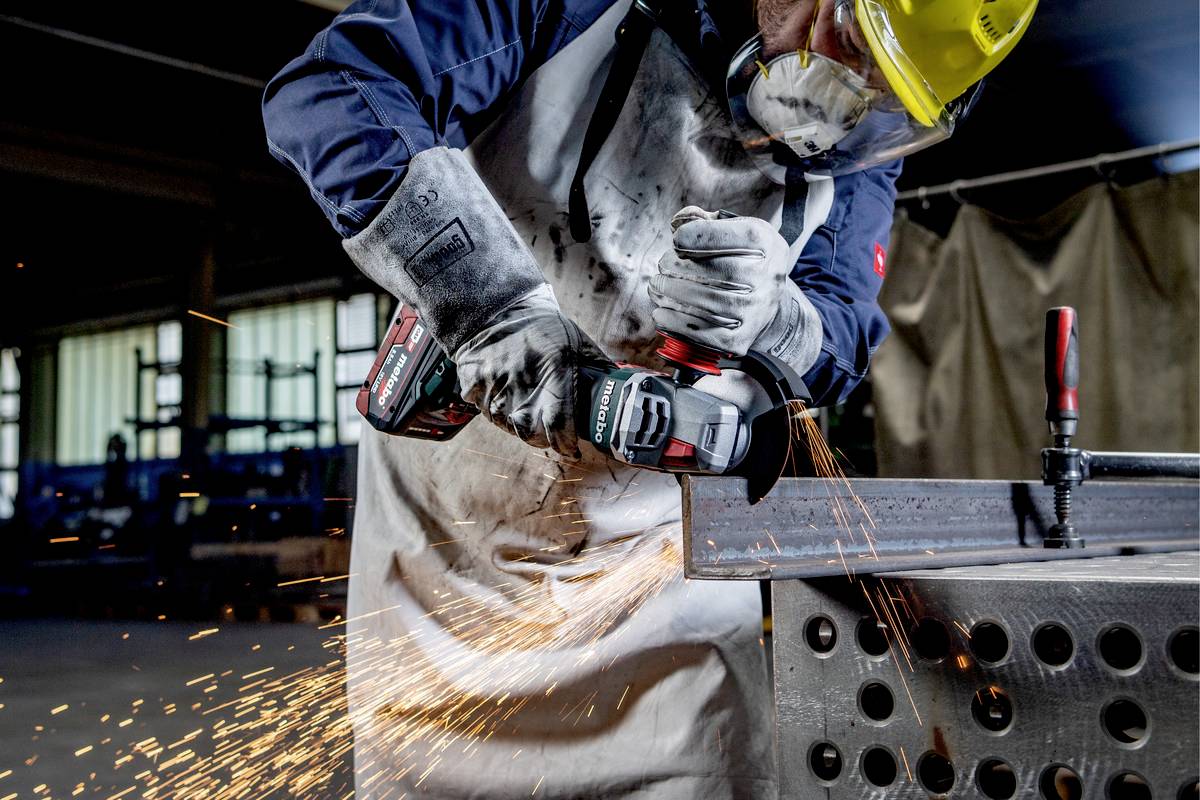 Osoba w odzieży ochronnej szlifuje metal szlifierką kątową, a wokół lecą iskry. W tle widoczny warsztat z warsztatowym stołem metalowym.