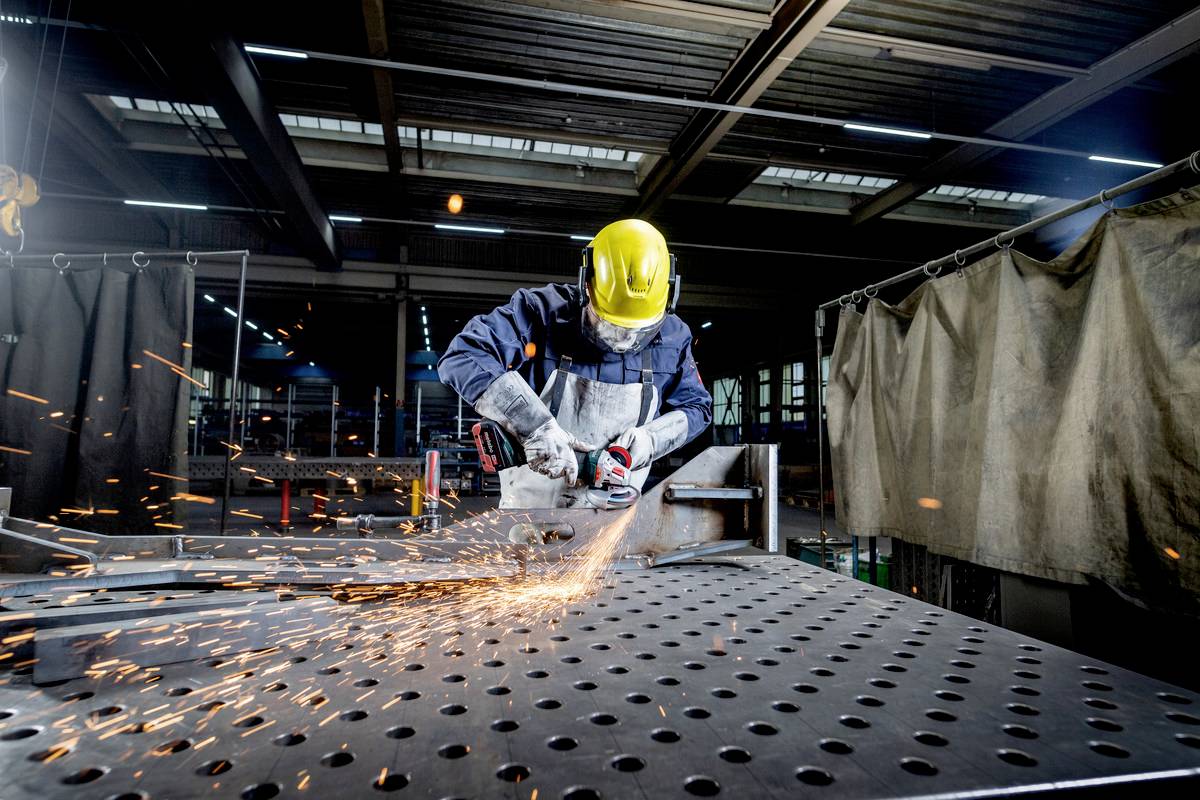 Osoba w odzieży ochronnej szlifuje metal w warsztacie. Iskry lecą z tarczy szlifierskiej podczas obróbki metalu.