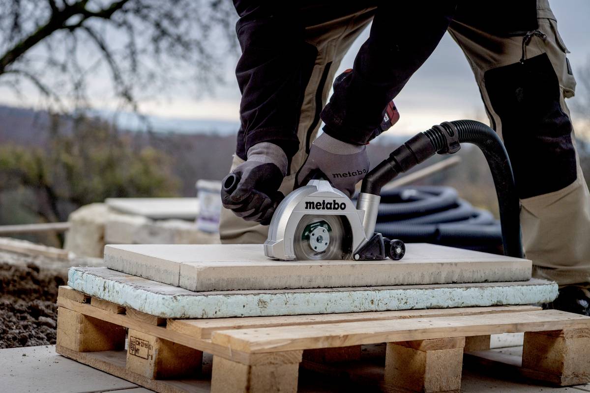 Robotnik kroi płytkę kamienną na budowie za pomocą elektrycznej piły. Widoczne są rękawice ochronne, odzież robocza oraz przyroda w tle.