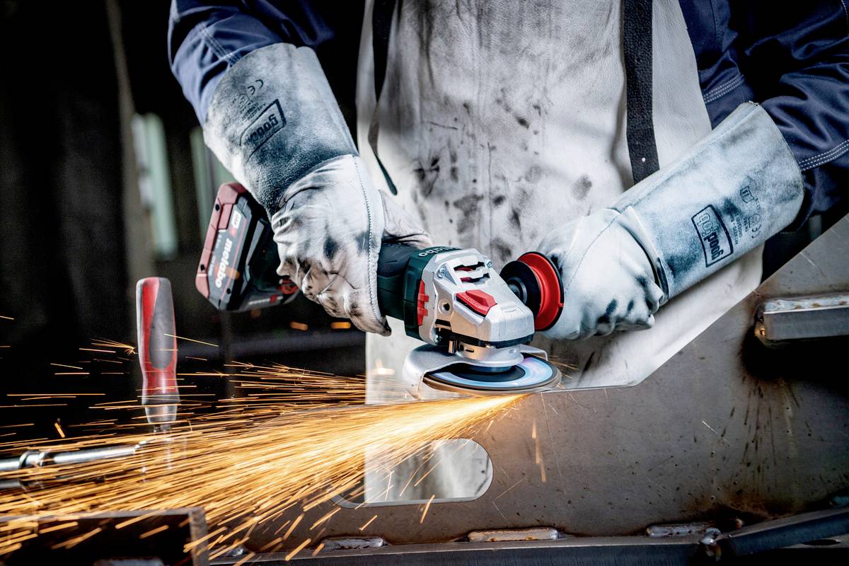 Osoba w sprzęcie ochronnym szlifuje metal elektronarzędziem, a wokół tryskają iskry. Focus na środkach bezpieczeństwa i precyzji.