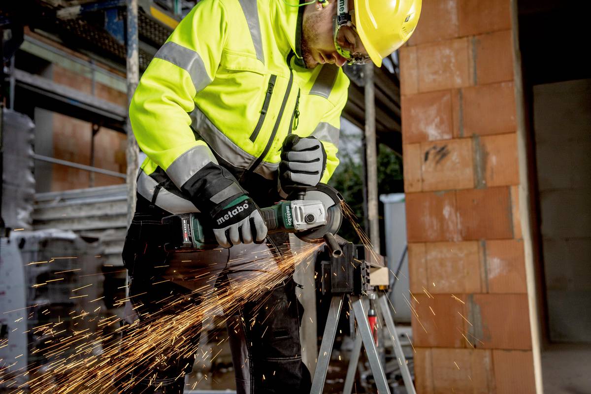 Robotnik budowlany w żółtym stroju ochronnym szlifuje metal kątówką, a wokoło lecą iskry. W tle widać plac budowy.