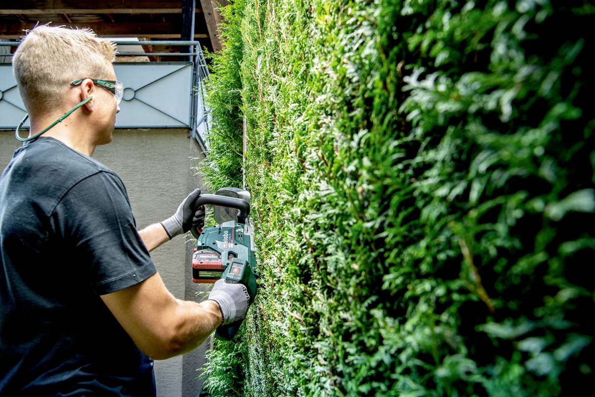 Mężczyzna przycina żywopłot elektrycznymi nożycami do żywopłotu. Nosi sprzęt ochronny, w tym okulary ochronne i rękawice.