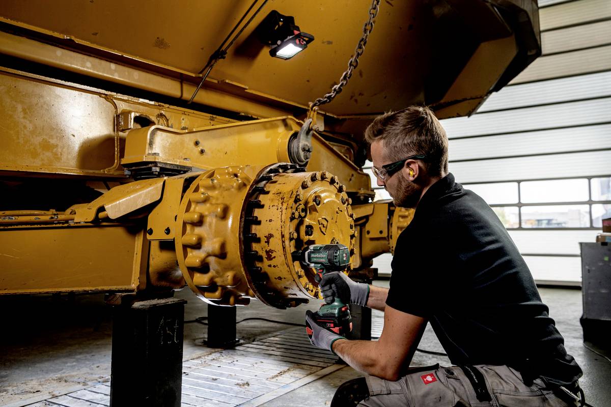 Mężczyzna w odzieży roboczej używa elektronarzędzia do pracy przy dużej żółtej części maszyny w warunkach warsztatowych.