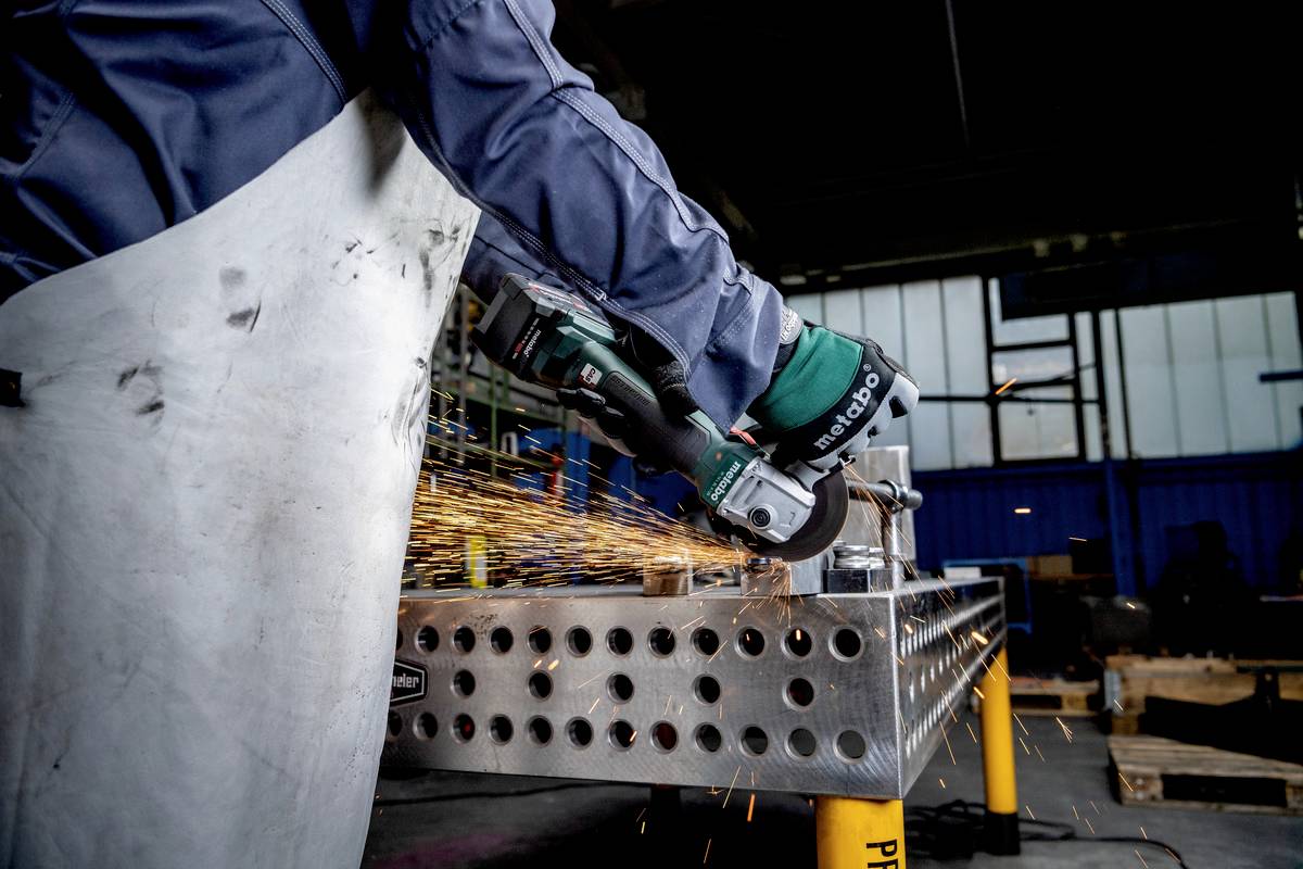 Osoba ubrana w sprzęt ochronny szlifuje metal szlifierką kątową, podczas gdy lecą iskry. Scena pracy w warsztacie.