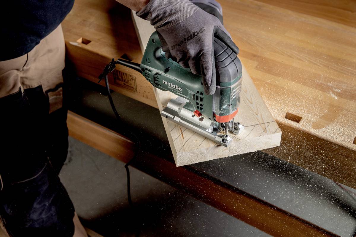 Osoba ucina kawałek drewna wyrzynarką w warsztacie. Wióry są widoczne, co wskazuje na aktywnie wykonywaną pracę.