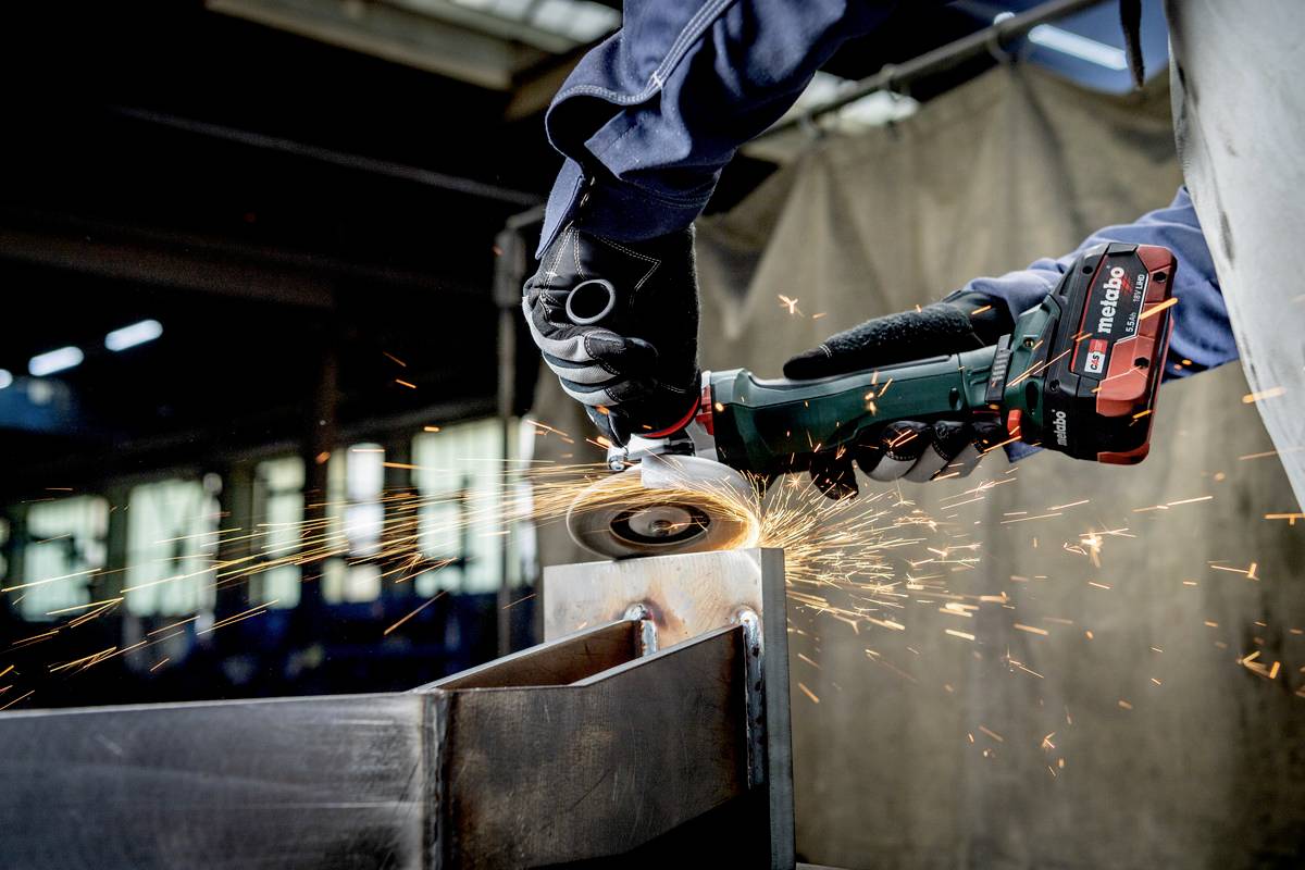 Osoba szlifuje metal kątrówką. Iskry lecą we wszystkich kierunkach. W tle widać wnętrze warsztatu.