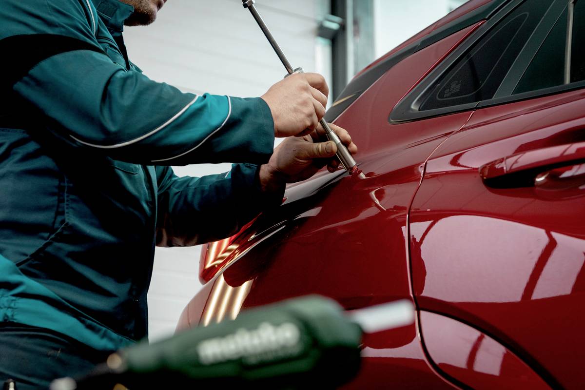 Osoba naprawia nadwozie czerwonego samochodu w warsztacie. Używa narzędzia, aby usunąć wgniecionę.