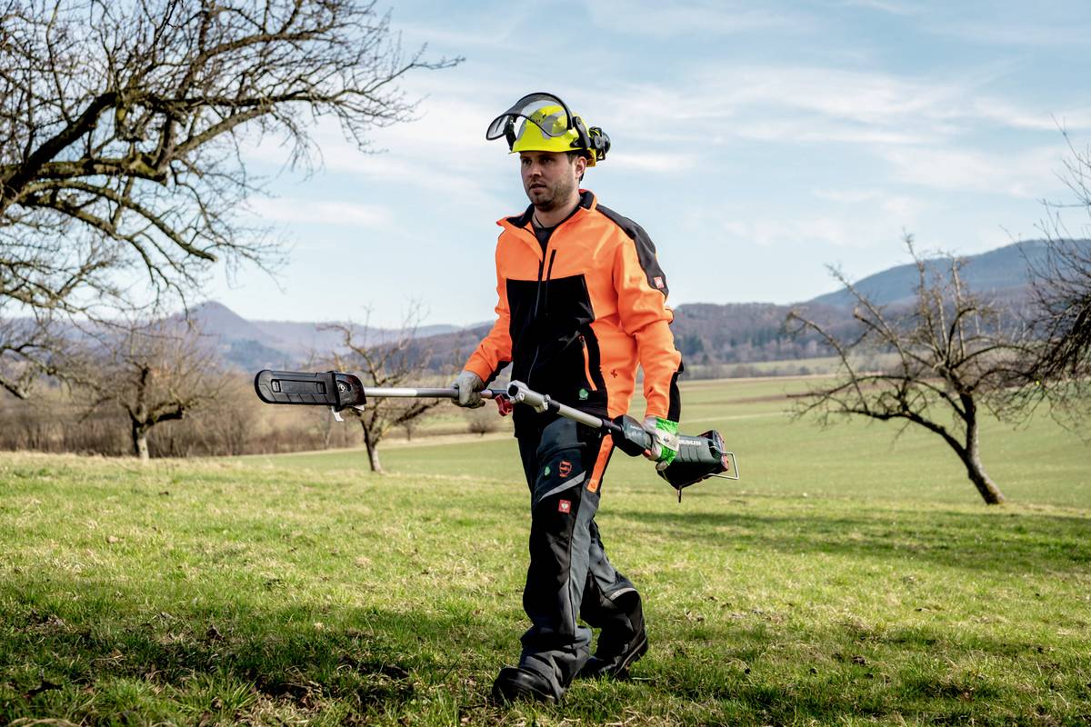 Mężczyzna w odzieży ochronnej kosi trawę na łące za pomocą podkaszarki. W tle widać nagie drzewa i góry.