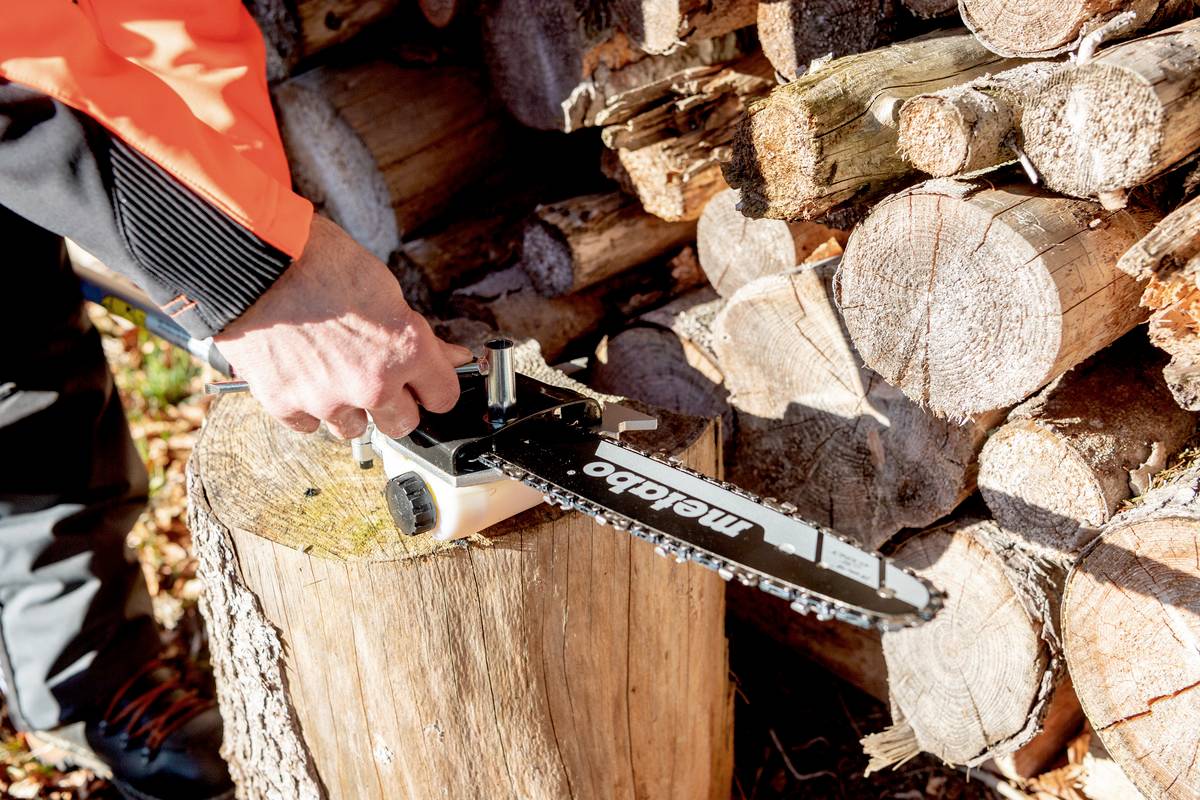 Osoba ostrzącza piłę łańcuchową na pniu drzewa przed stosem drewna.