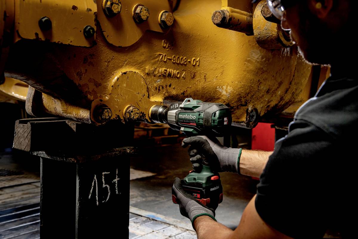 Osoba używa zielonego akumulatorowego wkrętarki do dokręcania śrub przy dużym, żółtym elemencie maszyny w warsztacie.