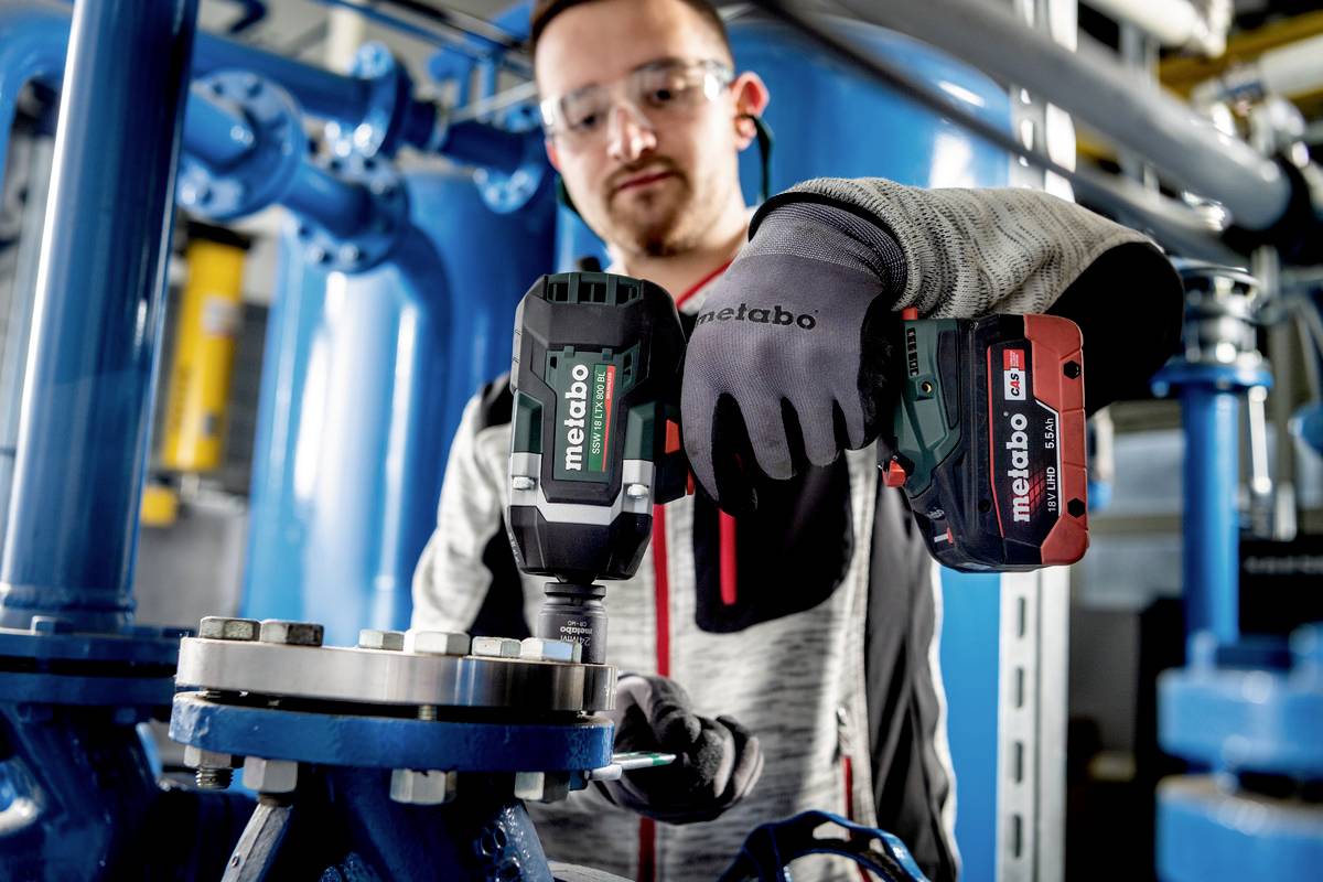 Mężczyzna w stroju roboczym używa akumulatorowego narzędzia Metabo do dokręcenia śruby przy maszynie przemysłowej.