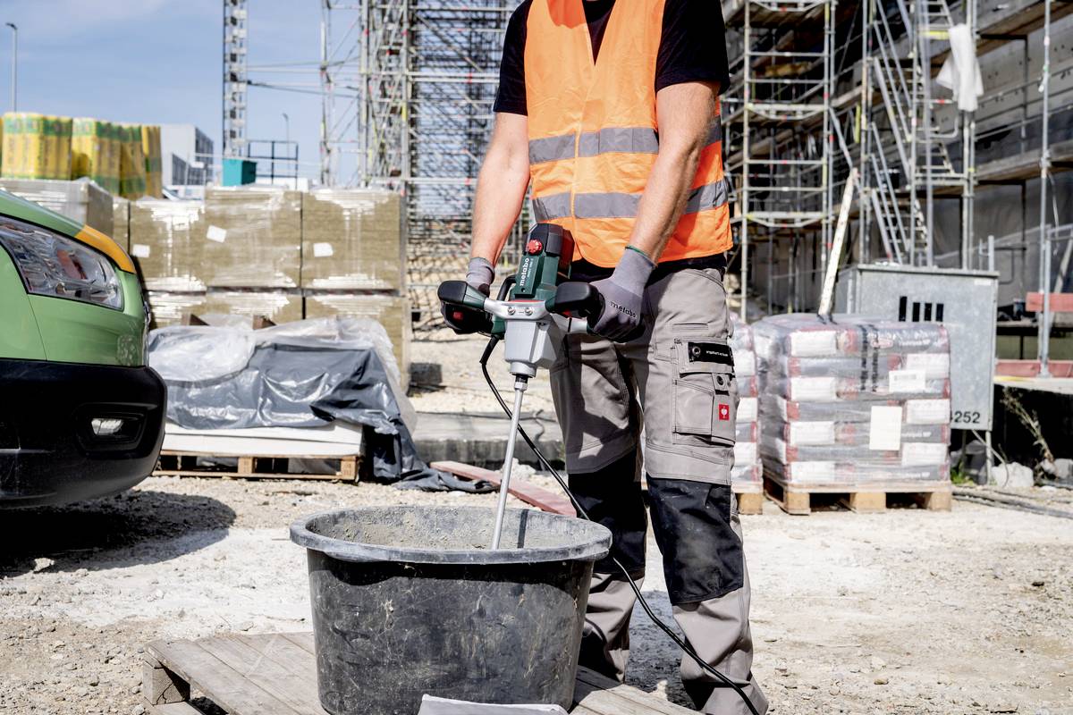 Robotnik budowlany w pomarańczowej kamizelce odblaskowej miesza cement w wiadrze na placu budowy. W tle widoczne są rusztowania i materiały budowlane.