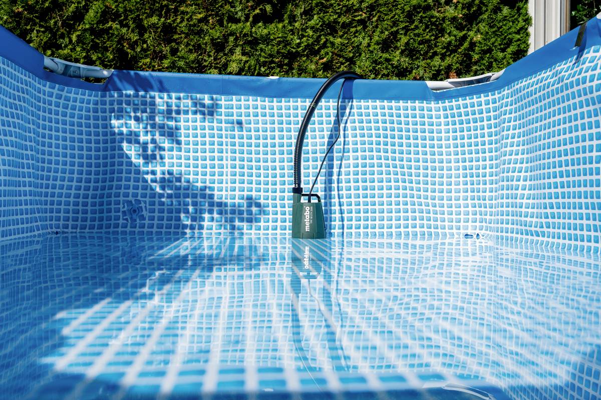 Pusty, dmuchany basen w niebiesko-białą kratkę. Pompka zwisa przez krawędź. W tle widoczne są zielone krzewy.