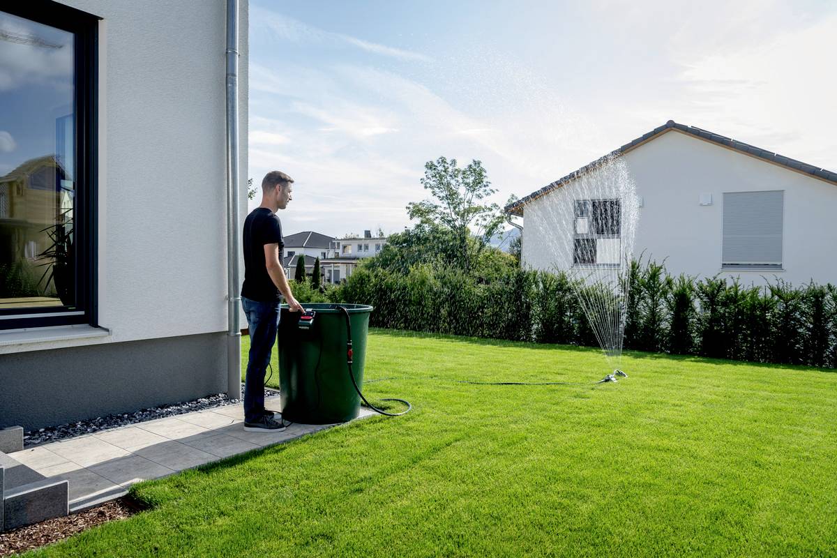Mężczyzna podlewa zielony trawnik za pomocą zraszacza. Obok niego stoi duży zielony pojemnik. W tle widać domy.
