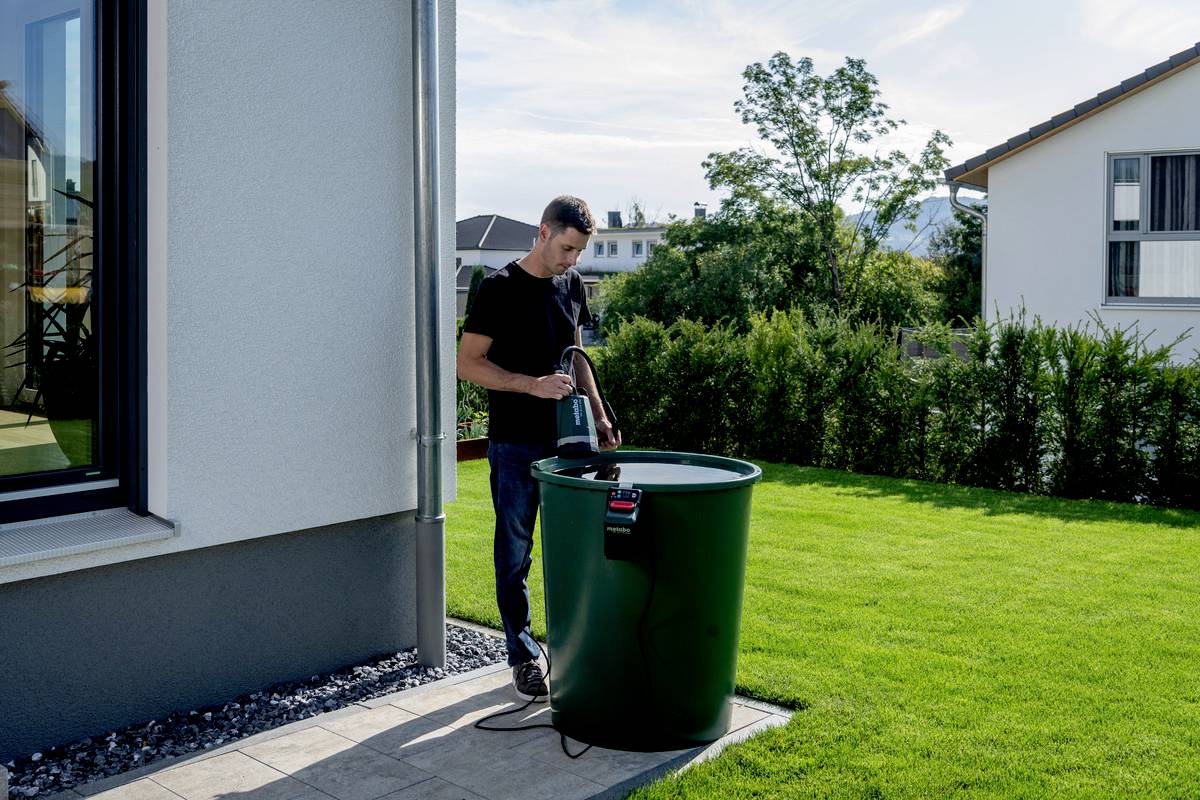Osoba napełnia wężem wodą duży, zielony pojemnik w ogrodzie obok domu. W tle widoczne są domy i drzewa.