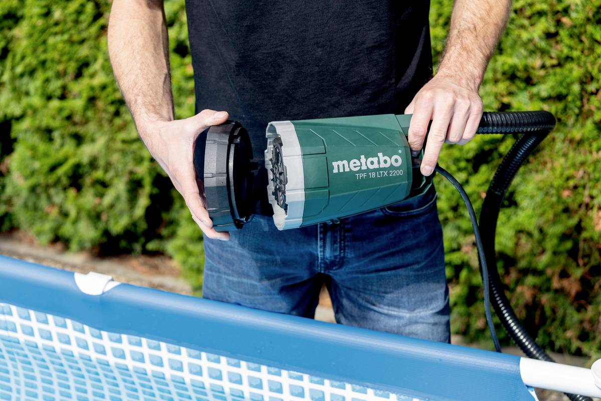 Osoba trzyma zieloną pompę wodną z napisem 'metabo', przygotowaną do użycia przy dmuchanym basenie przed żywopłotem.