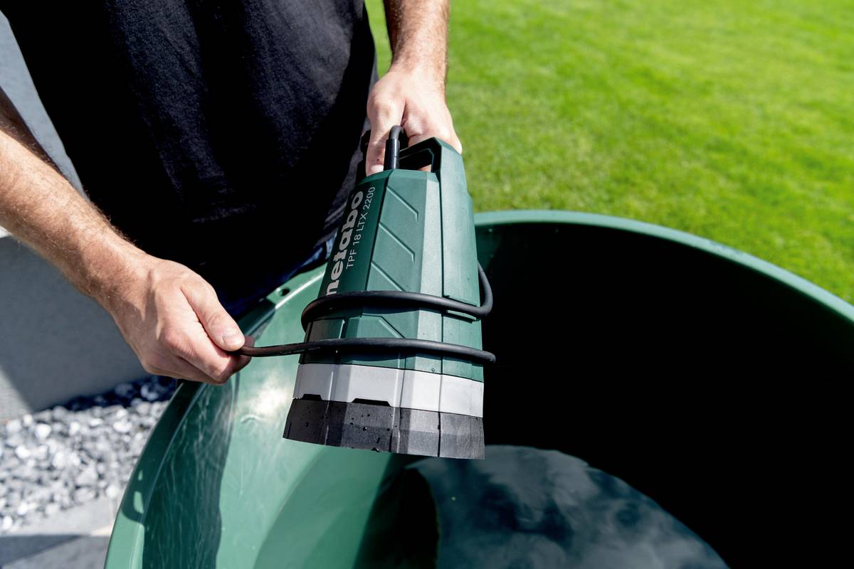 Osoba trzyma zieloną pompę wodną nad dużym wiadrem napełnionym do połowy wodą. W tle widać trawnik.