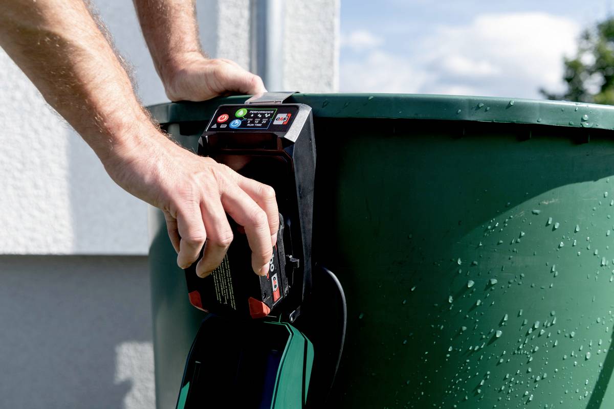 Osoba umieszcza akumulator na zielonym zbiorniku na deszczówkę na wolnym powietrzu. Akumulator wyświetla wskaźniki pracy, a obsługa jest wykonywana.