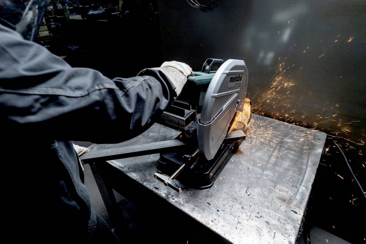Osoba przecina metalową rurę piłą metalową, podczas gdy tryskają iskry. Jest ubrana w odzież ochronną i rękawice.