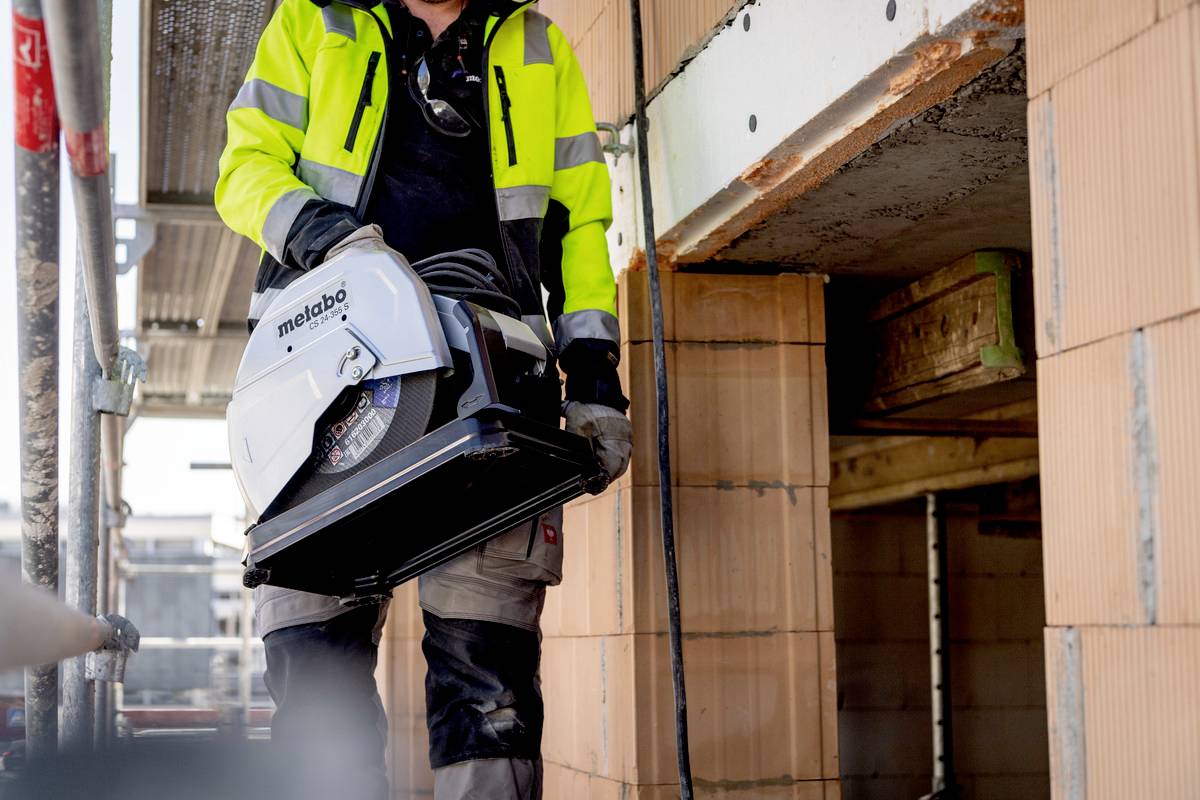 Robotnik budowlany w żółtej kamizelce odblaskowej trzyma ręczną piłę tarczową na placu budowy. W tle widoczne są niedokończone ściany.