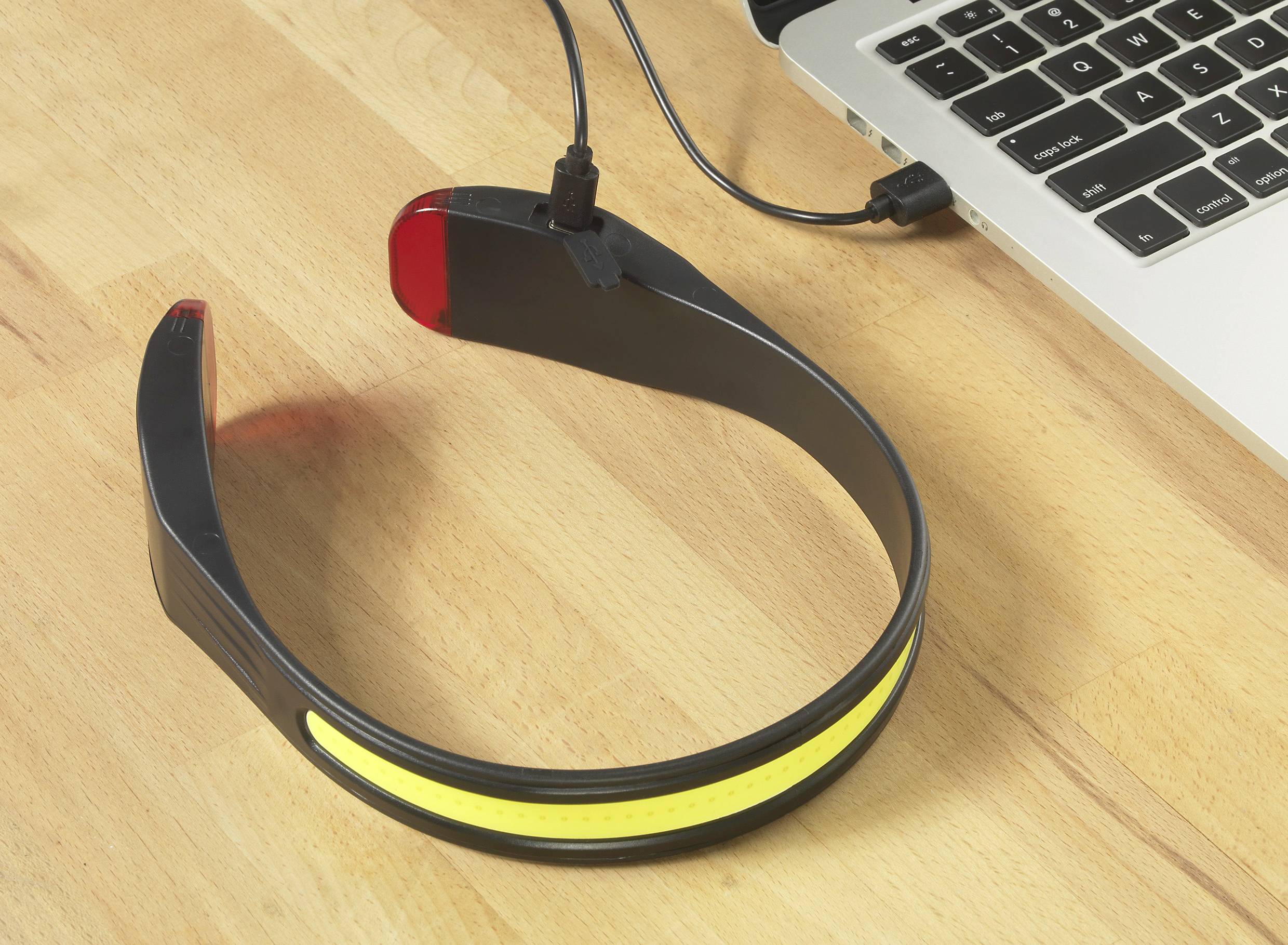 Czarna opaska na głowę z kablem USB leży obok laptopa na drewnianym stole. Opaska posiada żółty element świecący.