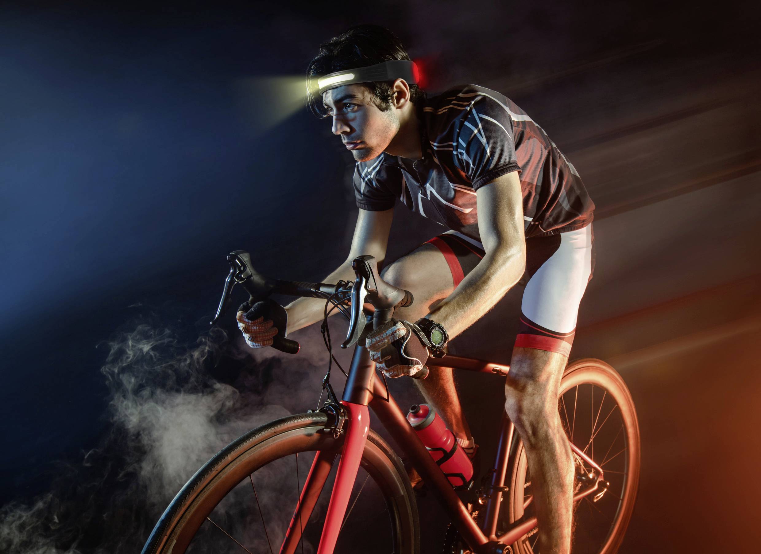 Rowerzysta w stroju sportowym intensywnie pedałuje, oświetlony czołówką, otoczony mgłą.