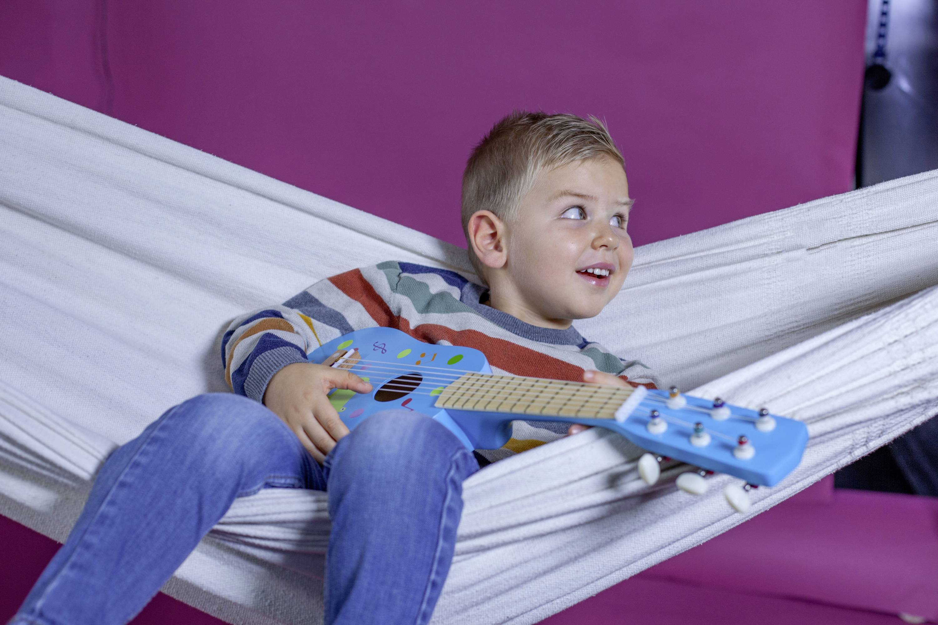 Chłopiec uśmiecha się, leżąc w białym hamaku na tle fioletowego tła, trzymając niebieską gitarę zabawkę.