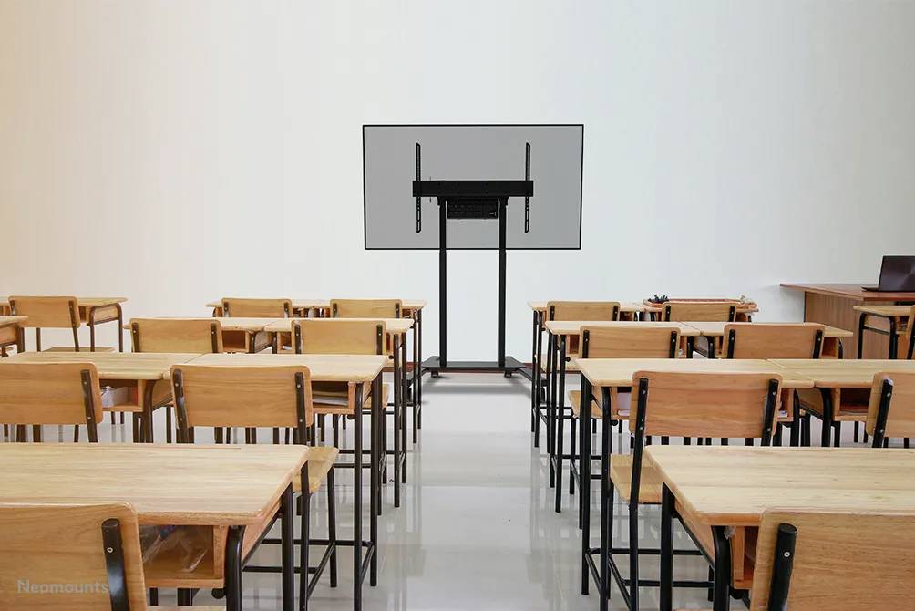 Sala lekcyjna z pustymi drewnianymi stolikami i krzesłami. Z przodu duży ekran. Po lewej i prawej stronie widoczne są ściany i biurko nauczyciela.