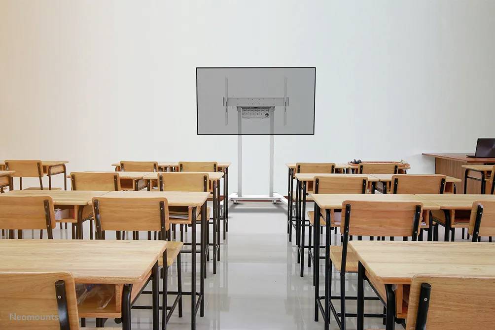 Sala lekcyjna z pustymi drewnianymi krzesłami i stolikami, dużym wyświetlaczem z przodu i biurkiem nauczyciela po prawej stronie obrazu.