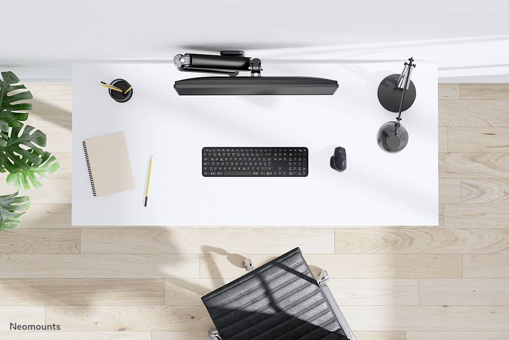 Schludnie urządzony biurowy blat z klawiaturą, myszką, lampką i notatnikiem, obok roślina i czarny fotel biurowy.