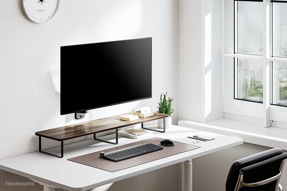 Minimalistyczne biurko z monitorem na drewnianym stojaku, klawiaturą, myszką, dokumentami i małą rośliną, tuż obok okna.