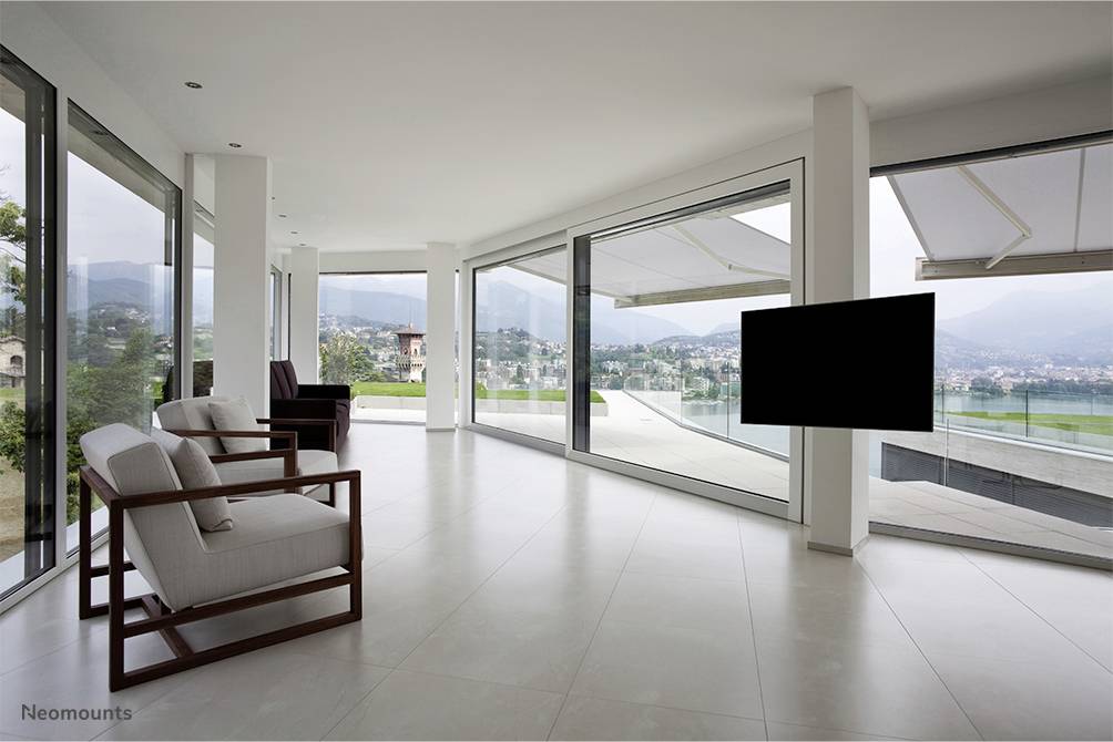 Nowoczesna, pusta przestrzeń mieszkalna z dużymi oknami i widokiem na rozległą okolicę. Po prawej stronie czarny telewizor jest zamontowany na ścianie.