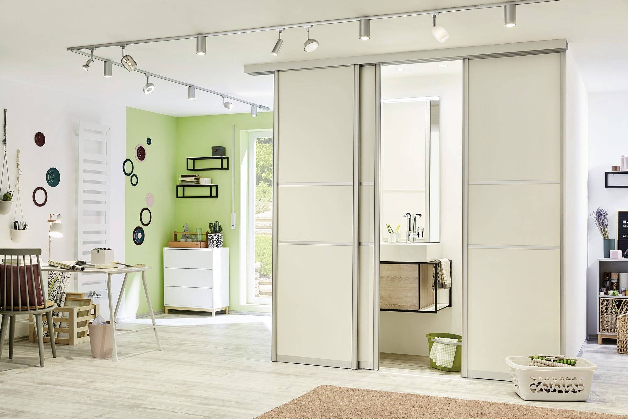 Jasny salon z wydzieloną strefą home office, biurkiem po lewej stronie, regałami przy zielonej ścianie, szafą z przesuwnymi drzwiami i listwą świetlną u góry.