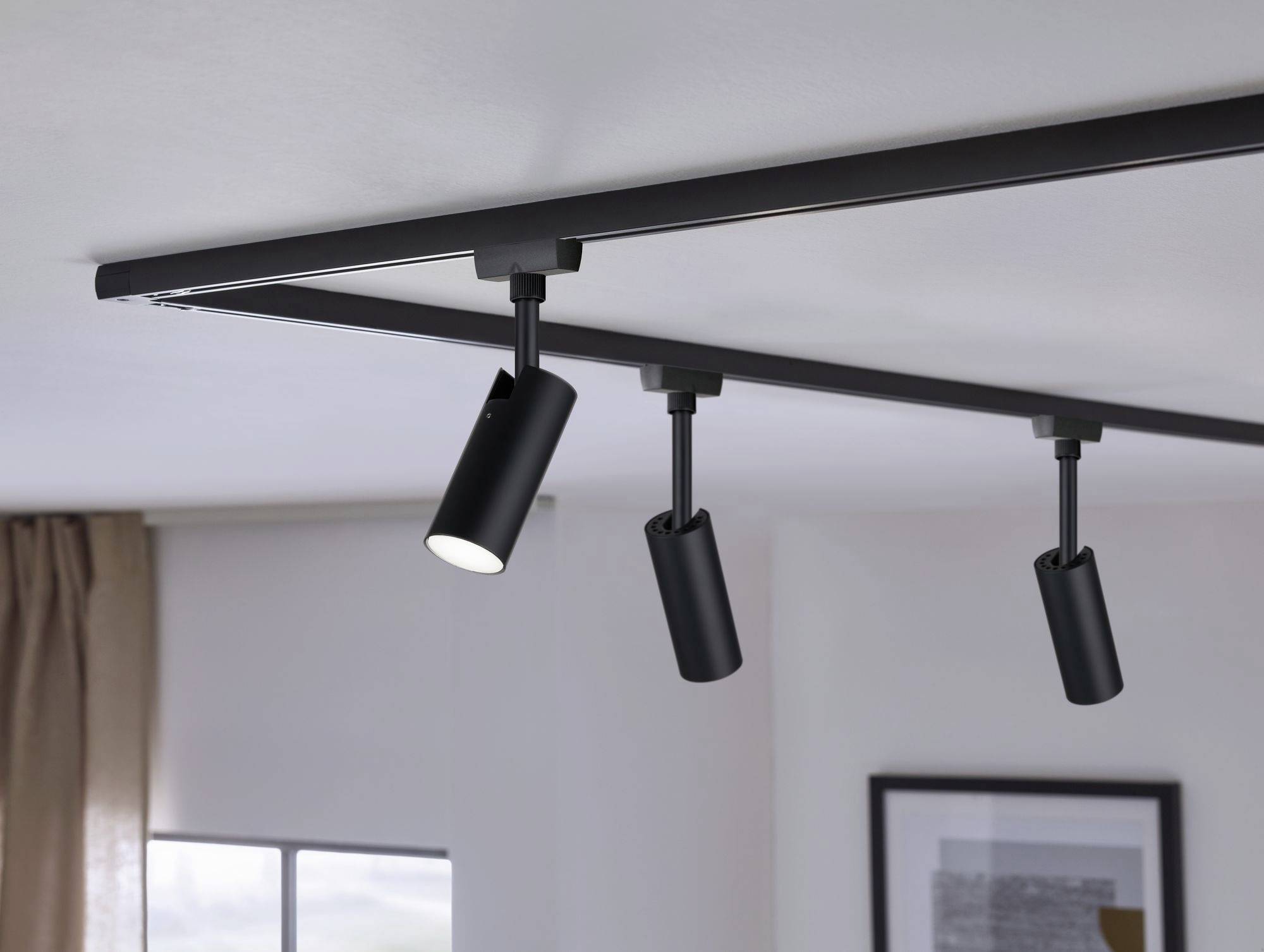 Trzy czarne reflektory sufitowe są zamontowane na szynie. Jeden z reflektorów jest włączony i świeci do pomieszczenia z oknami.