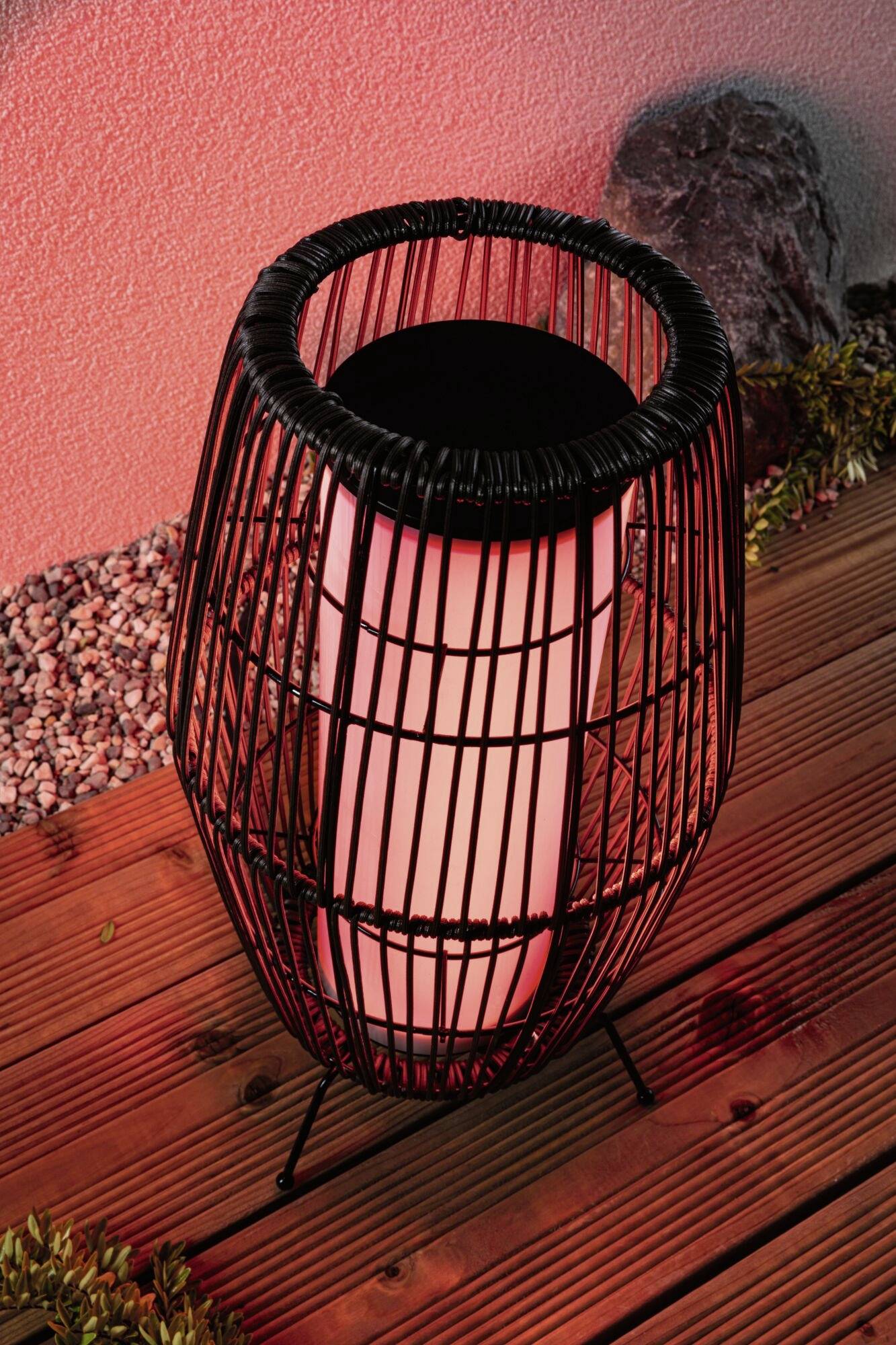 Latarnia Korb z czerwonym światłem na drewnianej podłodze, otoczona kamieniami i roślinami, tworzy nastrojowe oświetlenie.