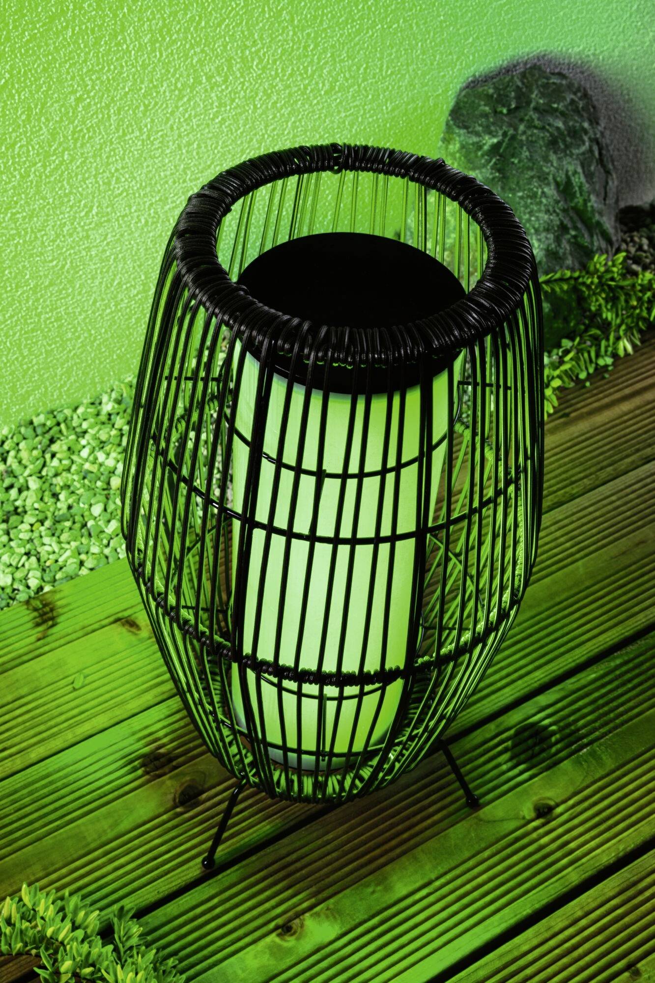 Czarna, okrągła latarnia z zielonym światłem stoi na drewnianym podłożu. W tle widoczne są zielone kamyczki i oświetlona ściana.
