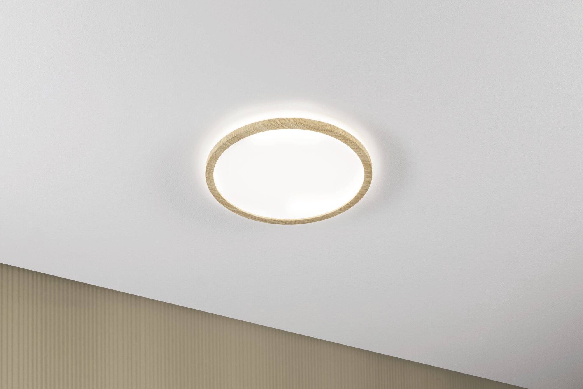 Okrągła, nowoczesna lampa sufitowa z drewnianą ramą, świecąca w neutralnym pomieszczeniu.