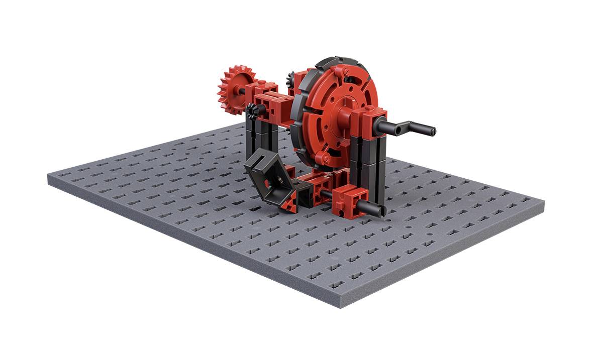 Mechaniczny model składający się z czerwonych i czarnych elementów na szarej, perforowanej podstawie. Prezentuje on koła zębate i dźwignie łączące poszczególne części konstrukcji.