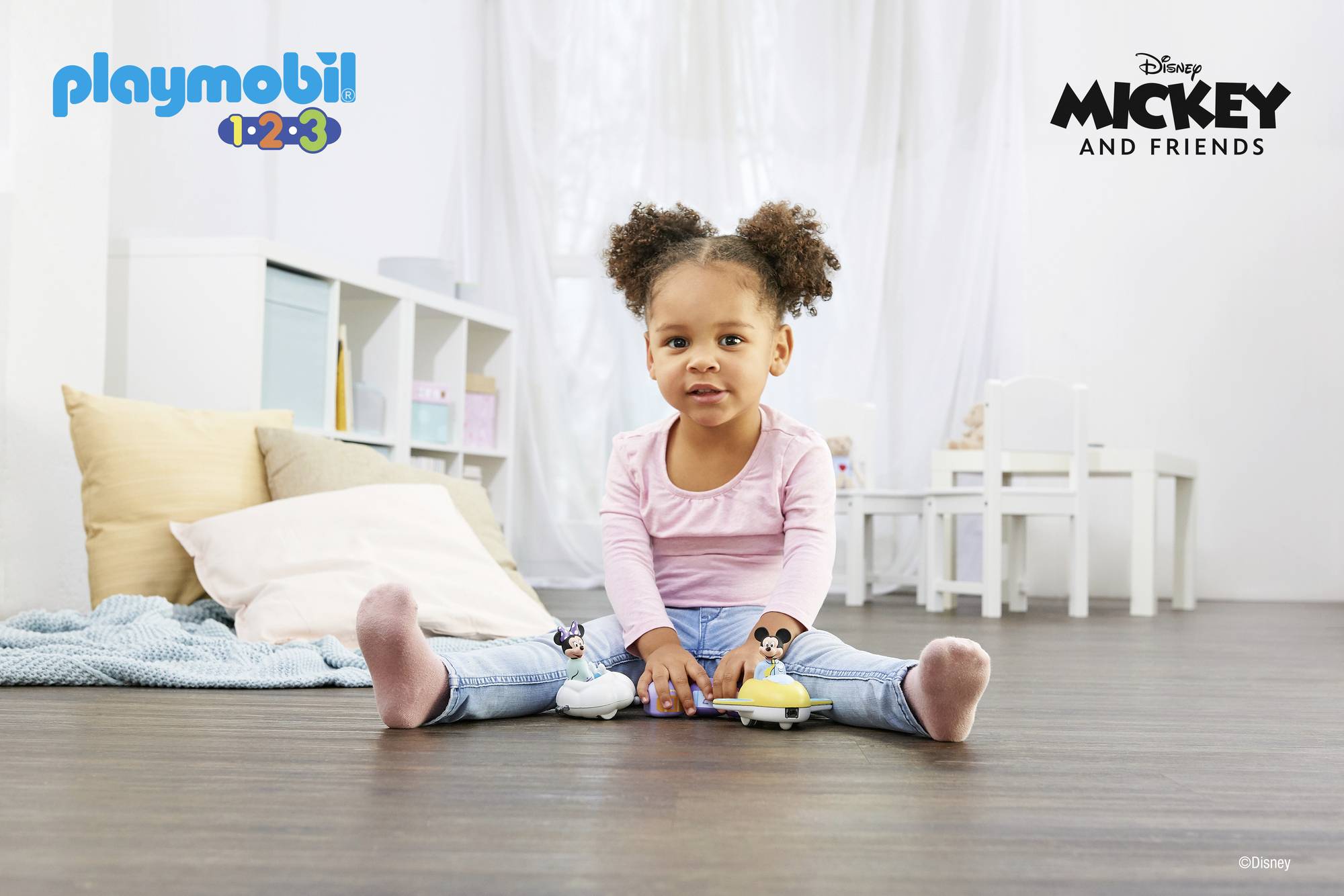 Małe dziecko siedzi na podłodze i bawi się dwoma zabawkami Myszki Miki. W tle widać regały i stół.