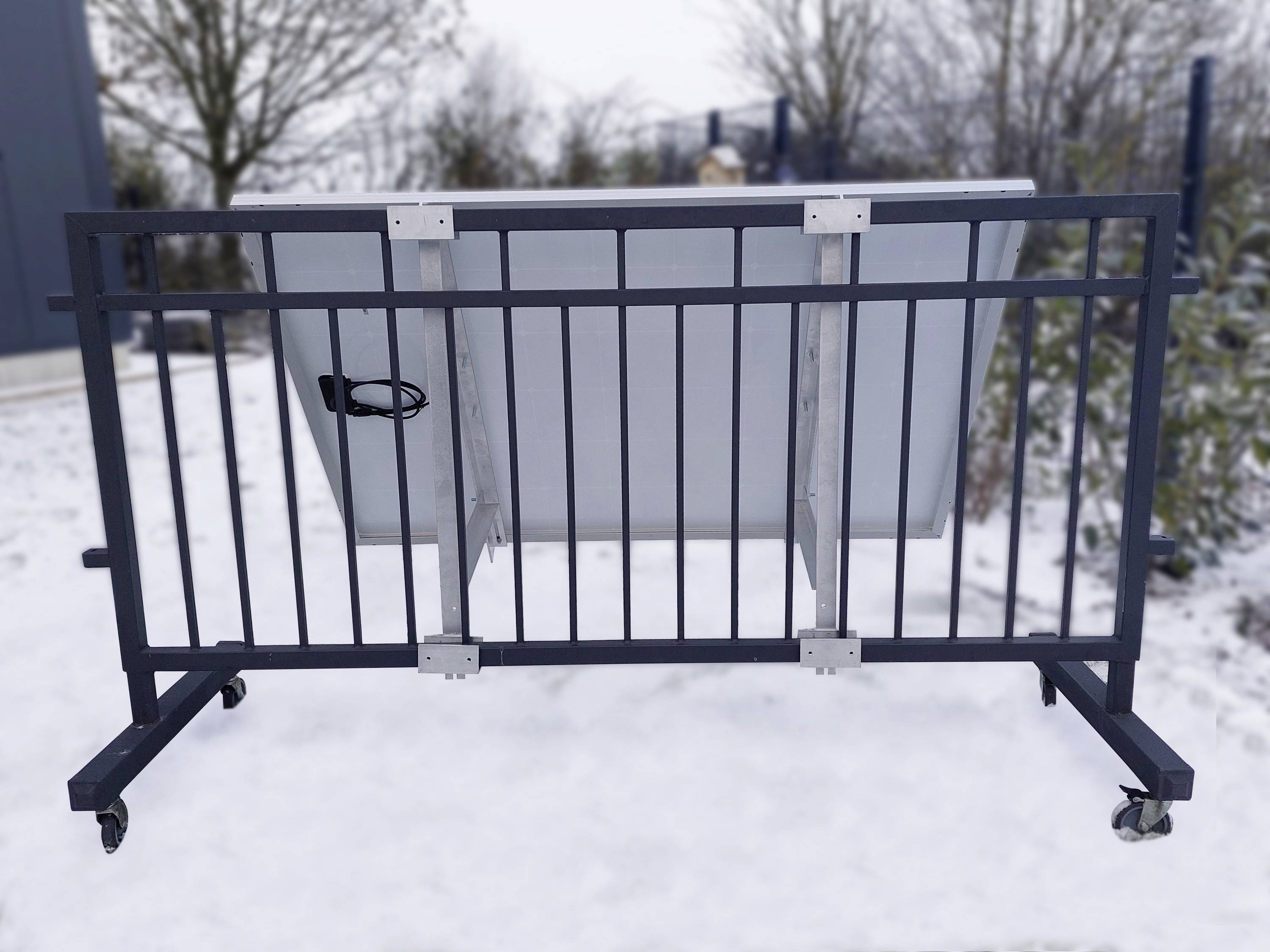 Mobilna bariera na dwóch kołach z przewodem z tyłu stoi na zaśnieżonej powierzchni. W tle widoczne są drzewa.